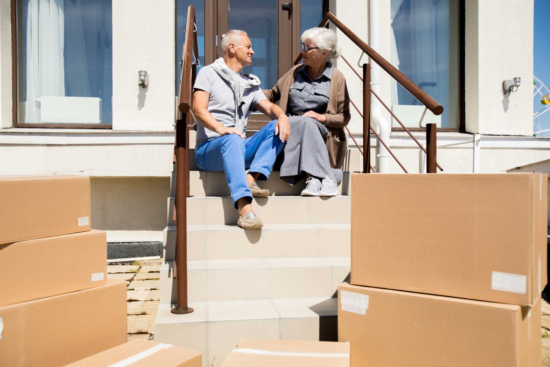 Two seniors smiling at each other sitting on their front stoop, surrounded by moving boxes.